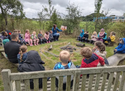 A group of children sit in a circle on logs around a small campfire pit as an adult reads a picture book outdoors under a cloudy sky.