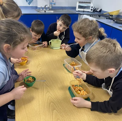 Children in aprons sit around a wooden table in a classroom kitchen, eating pasta from plastic containers with forks, blue cabinets and a sink visible in the background.