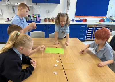 Several students in light blue shirts build structures from dry spaghetti and marshmallows at a classroom table, with a green sheet in the center.