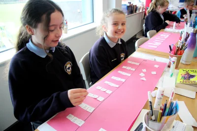 Two students sit at a craft table in a bright classroom; one smiles at the camera while arranging small cards on a strip with supplies nearby.