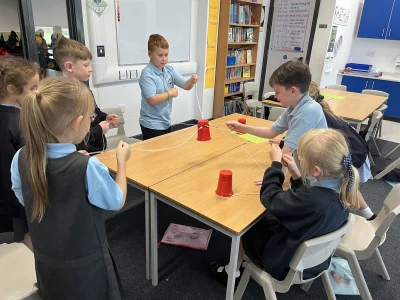 Group of schoolchildren working at a classroom table, threading string through red cups in a hands-on STEM activity.