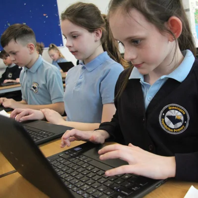 Students in a classroom using laptops; in the foreground, a student in a dark cardigan with a crest types on a laptop.