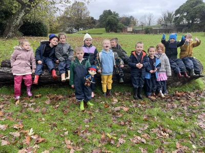 Group of kids in coats and rain boots sit on a fallen log in a park, autumn leaves on the ground, smiling and giving thumbs up.