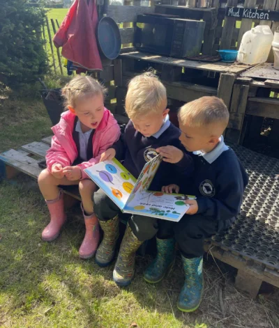 Three children in school uniforms sit on a pallet bench outdoors, reading a colorful picture book together, with boots and a mud kitchen nearby.