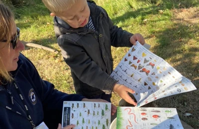 An adult staff member wearing a lanyard and a child look at a large illustrated butterfly and insect guide outdoors on grass.