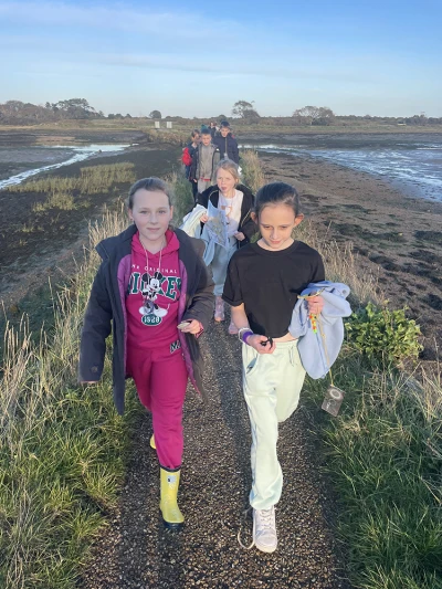 A group of children walks along a gravel path beside a tidal marsh, with grassy banks, distant trees, and a clear blue sky.