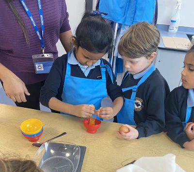 Three students in blue aprons and uniforms stand at a table cracking eggs into a small red bowl, supervised by an adult nearby.