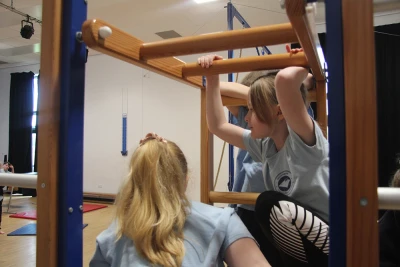 Several people in light blue shirts use a wooden gymnastic frame, gripping the bars inside a school gym with mats on the floor.