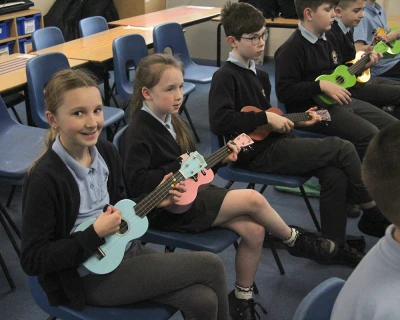 Group of schoolchildren in uniform sit on blue chairs in a classroom, each holding a pastel-colored ukulele and playing together.