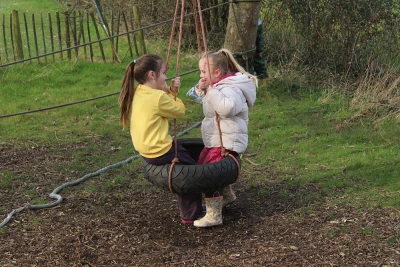 Two children on a tire swing facing each other, gripping the ropes in a grassy yard with a wooden fence behind them; one wears a yellow hoodie, the other a light jacket.